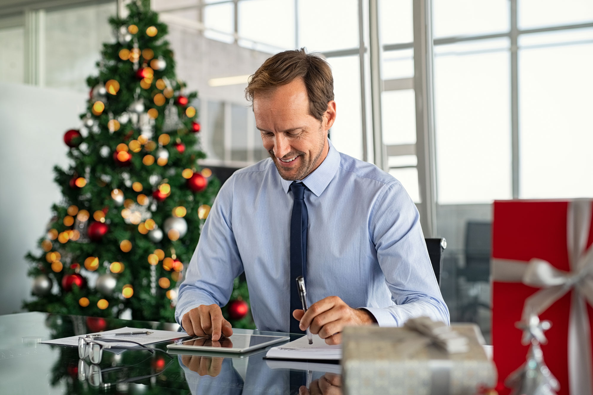 Business professional using a digital planner on a tablet at a festive office setting with a decorated Christmas tree and holiday gifts.