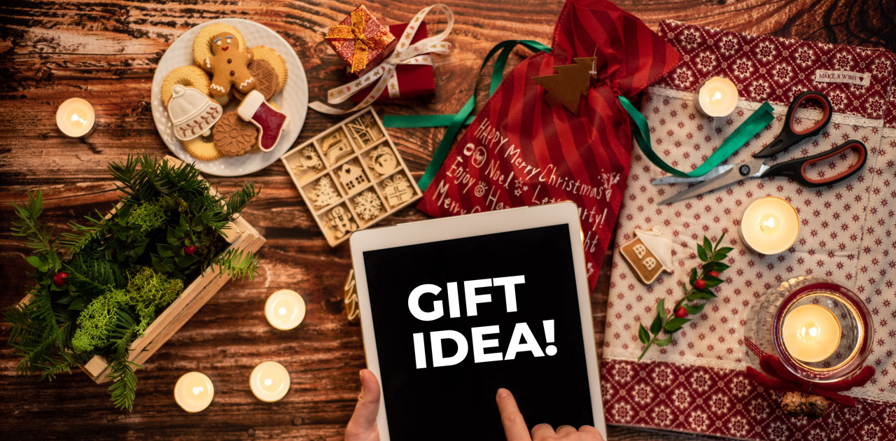 Hand holding tablet displaying "GIFT IDEA!" surrounded by holiday-themed decorations, cookies, candles, and gift wrapping materials, illustrating unique gift suggestions for employees and bosses.