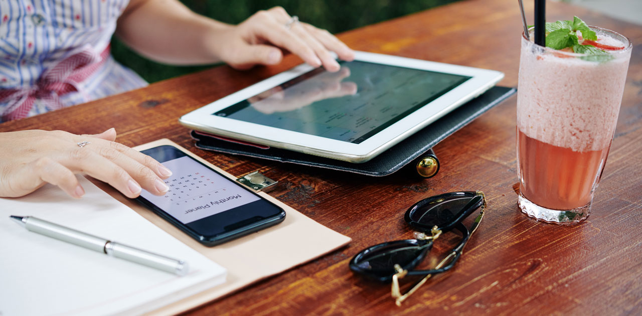 Person using a tablet and smartphone on a wooden table, with a notebook, pen, sunglasses, and a refreshing drink, illustrating digital planning and organization.
