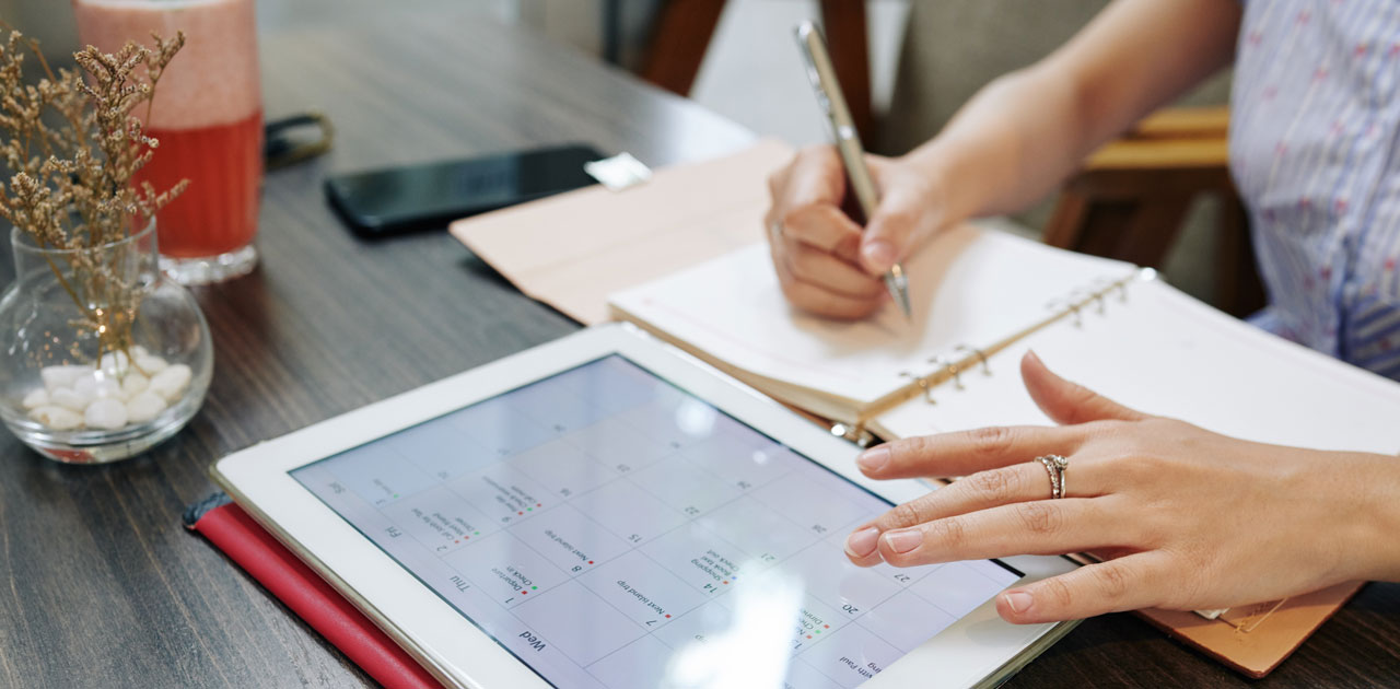 Person using digital planner on tablet, writing notes in notebook, with a drink and decorative plant in a workspace, illustrating strategies for effective digital planning.