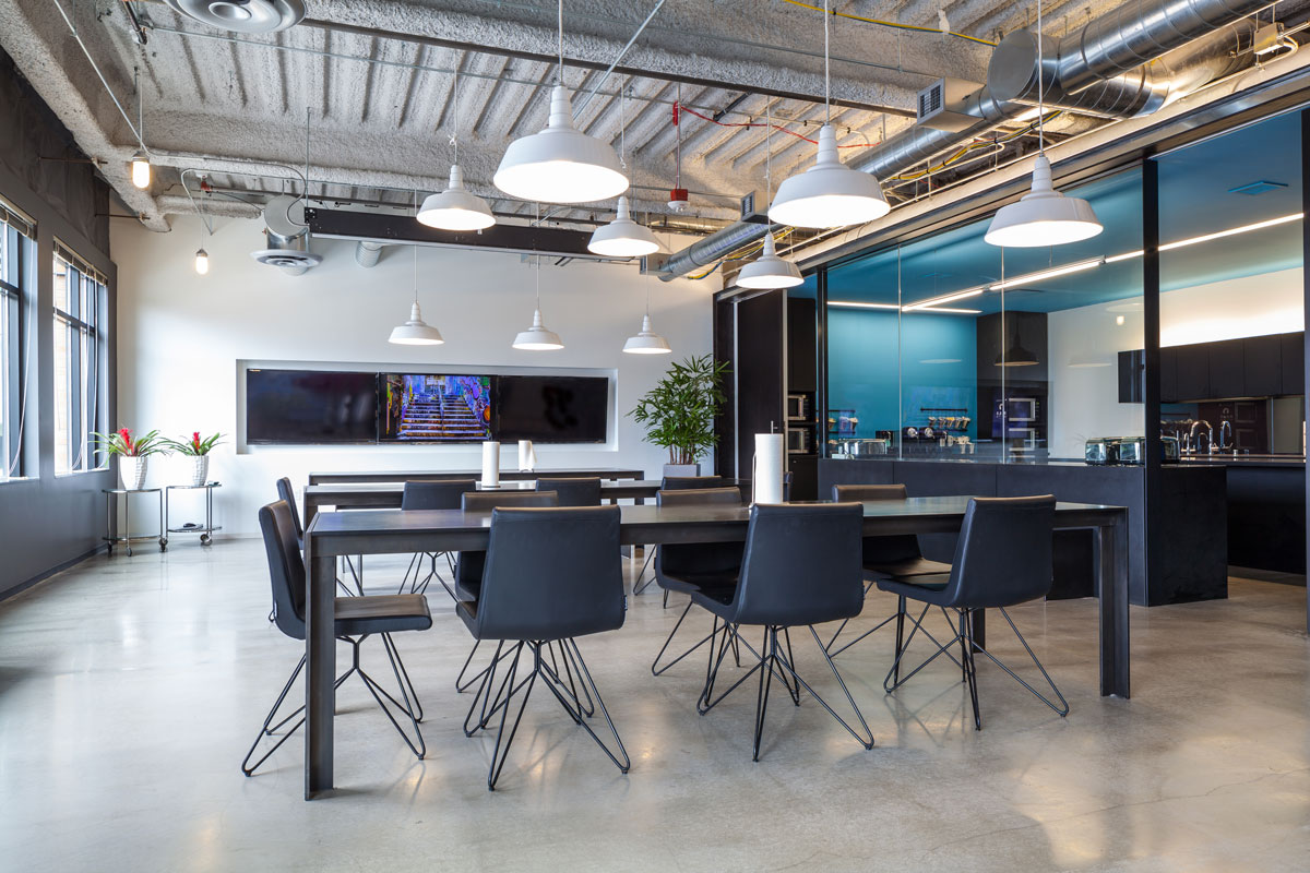 Open plan office dining area featuring modern tables and chairs, large windows, hanging lights, and a kitchenette in the background, illustrating collaborative workspace design.