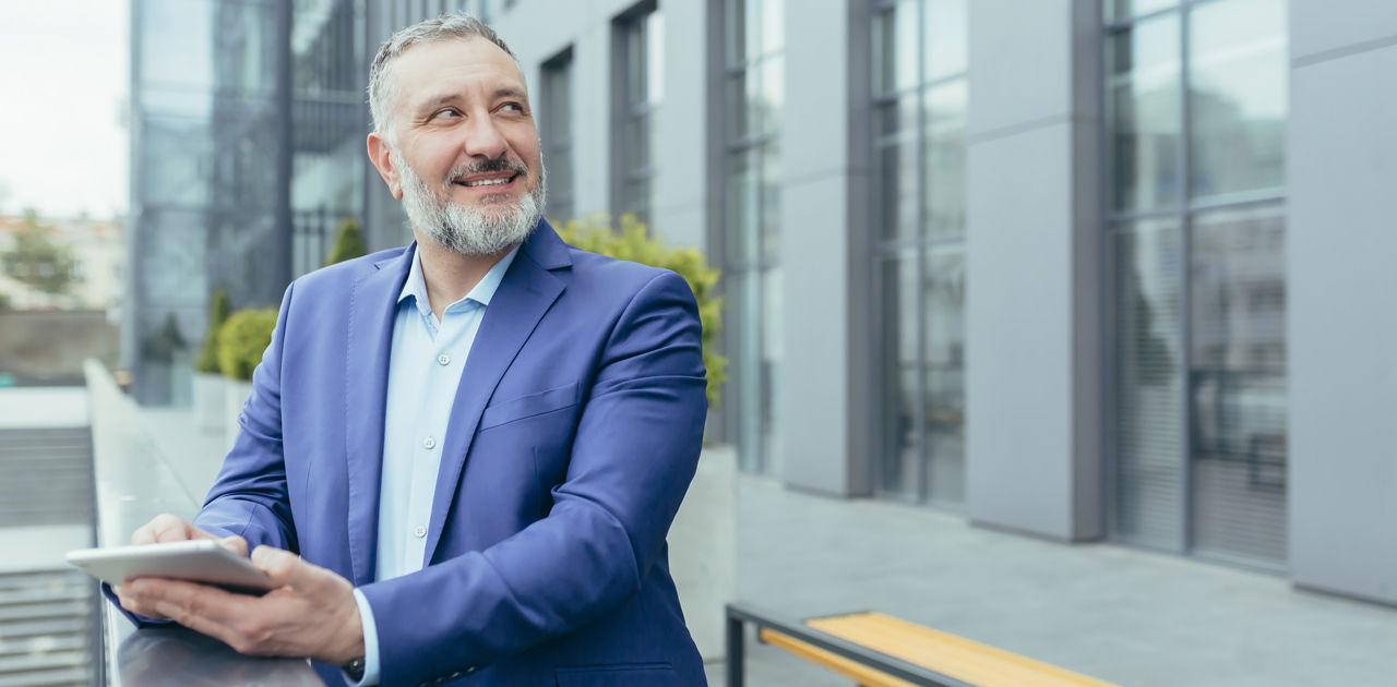 Smiling man in a business suit holding a digital tablet, representing productivity and technology for academic administrators and staff, with modern building backdrop.