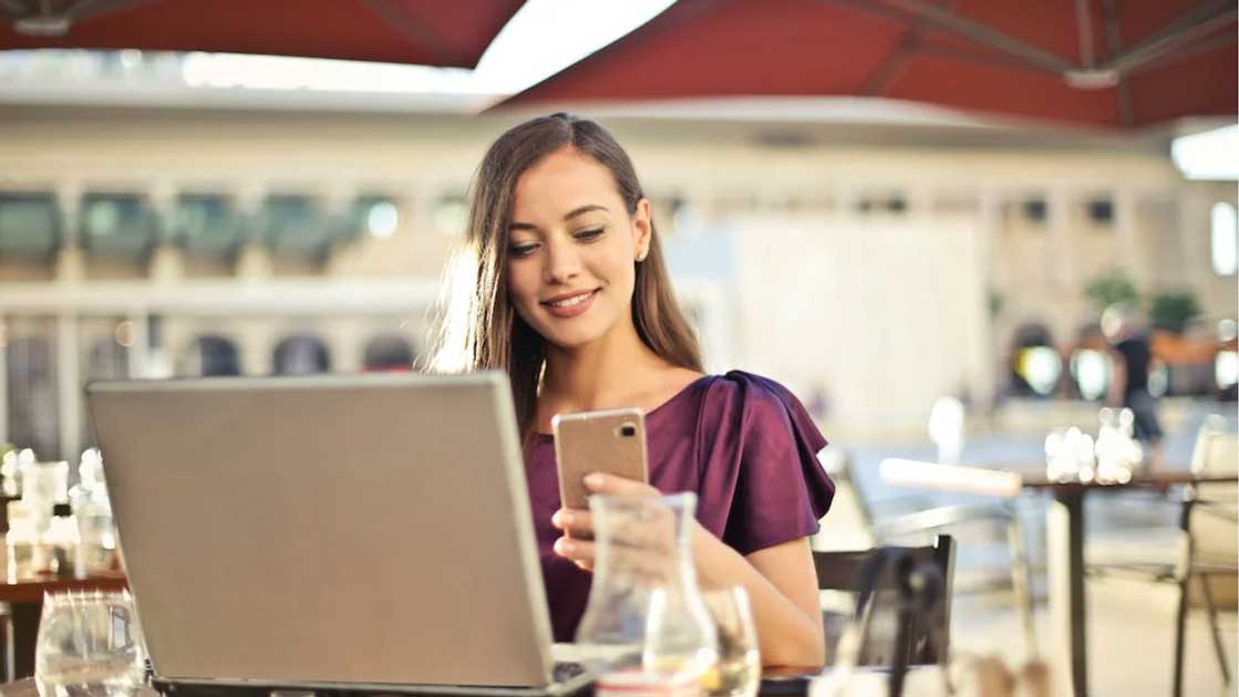 Woman smiling while using a smartphone and laptop at a café, representing the transition from solopreneur to business owner.