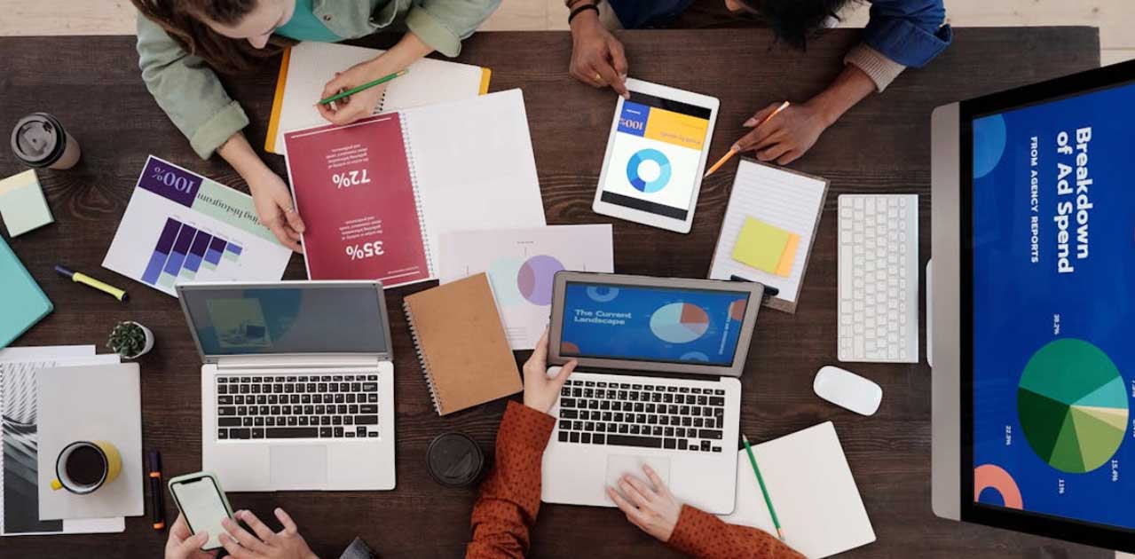 Group of professionals analyzing digital marketing data on laptops and tablets, featuring charts and graphs, with emphasis on ad spend breakdown and performance metrics.